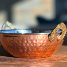 A close-up of a hammered copper kadhai pan with brass handles, resting on a wooden surface with a blurred background.