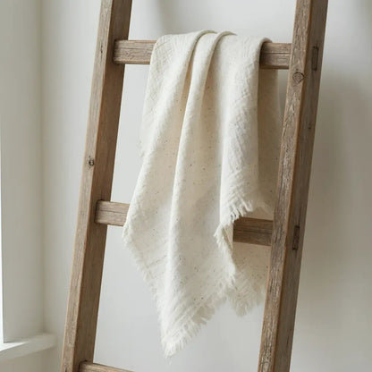 Cream-colored linen throw with subtle speckles and frayed edges draped over rustic ladder in muslin bath towel style