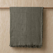 A soft olive-green lightweight handwoven cotton blanket with fringed edges hangs on a wooden rod
