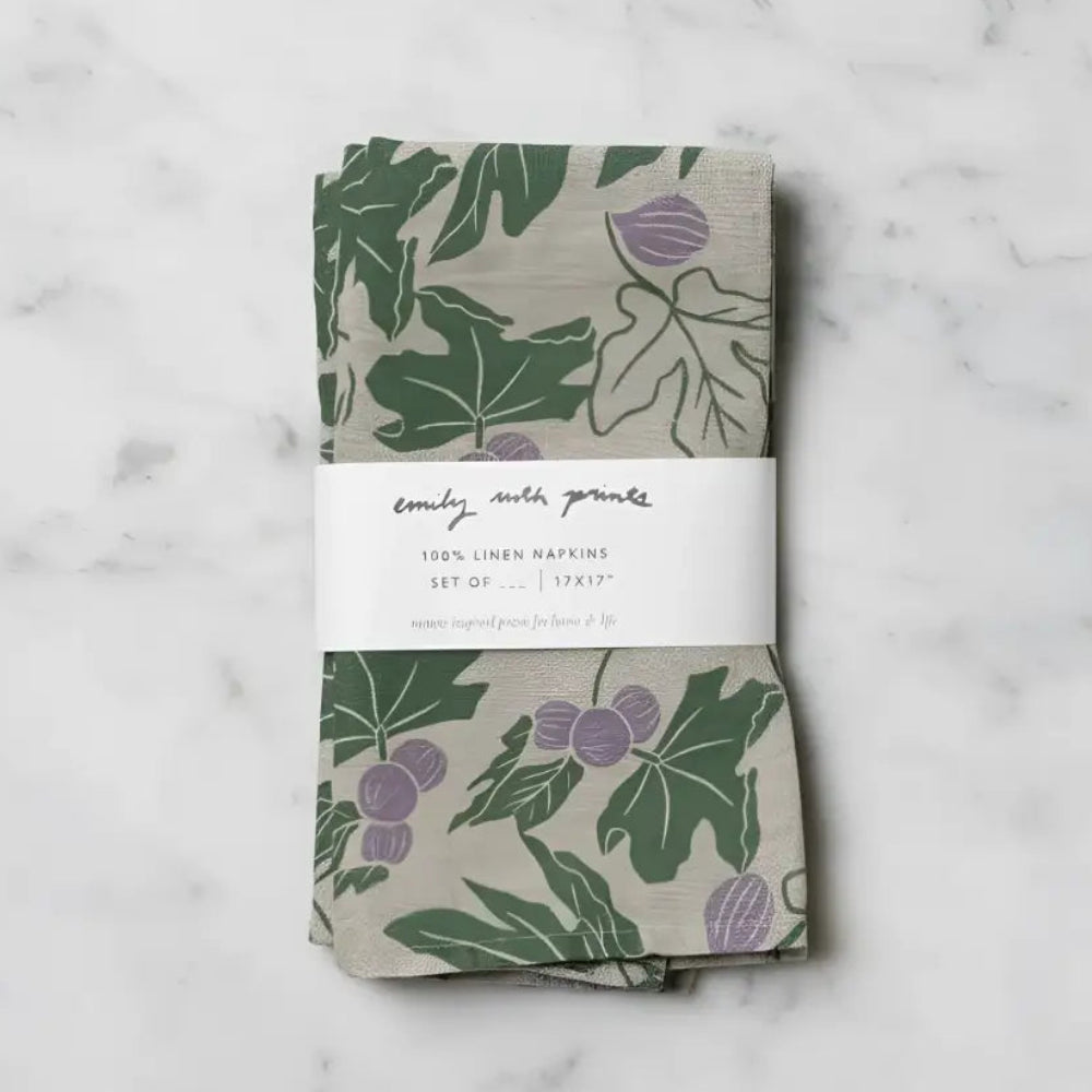 A set of two beige linen napkins with fig designs and green leaves wrapped in a white label band