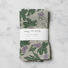 A set of two beige linen napkins with fig designs and green leaves wrapped in a white label band