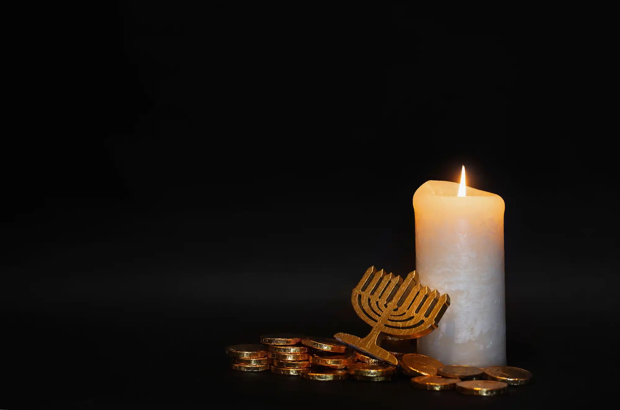 A lit white pillar candle next to a golden menorah and gold coins.