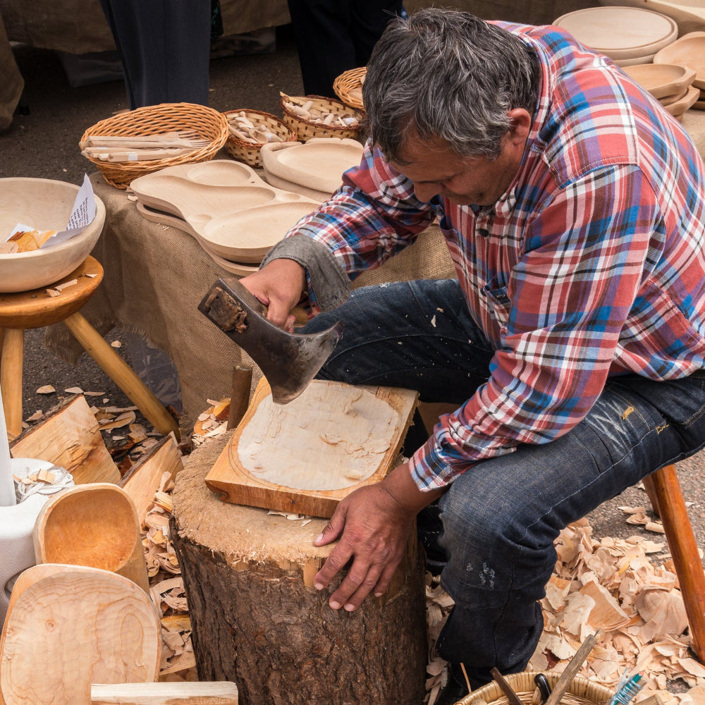 A man in a red and blue plaid shirt carves a wooden board using a hand tool on a log stump at an outdoor market, surrounded by wood scraps, bowls, and tools.