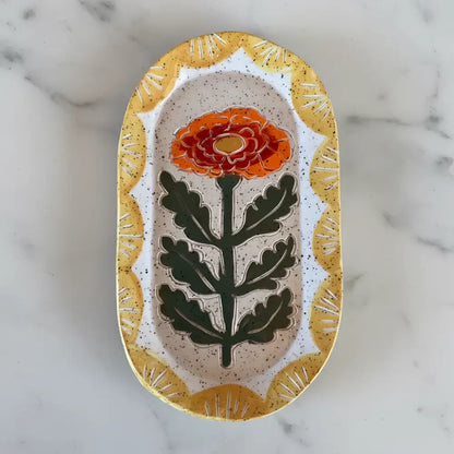 An oval handcrafted ceramic trinket dish with a marigold flower and golden-yellow border perfect for small items