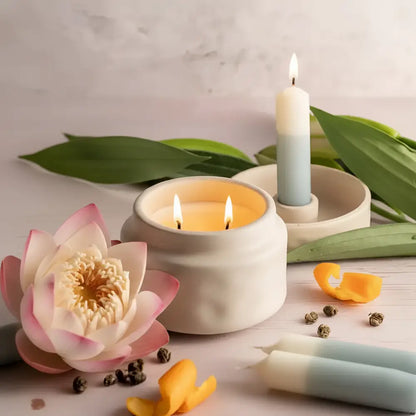 A white ceramic mindful moments candle jar with two lit candles surrounded by pink lotus and green leaves
