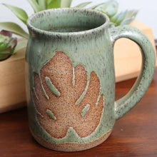 Speckled stoneware green mug with brown monstera leaf design.