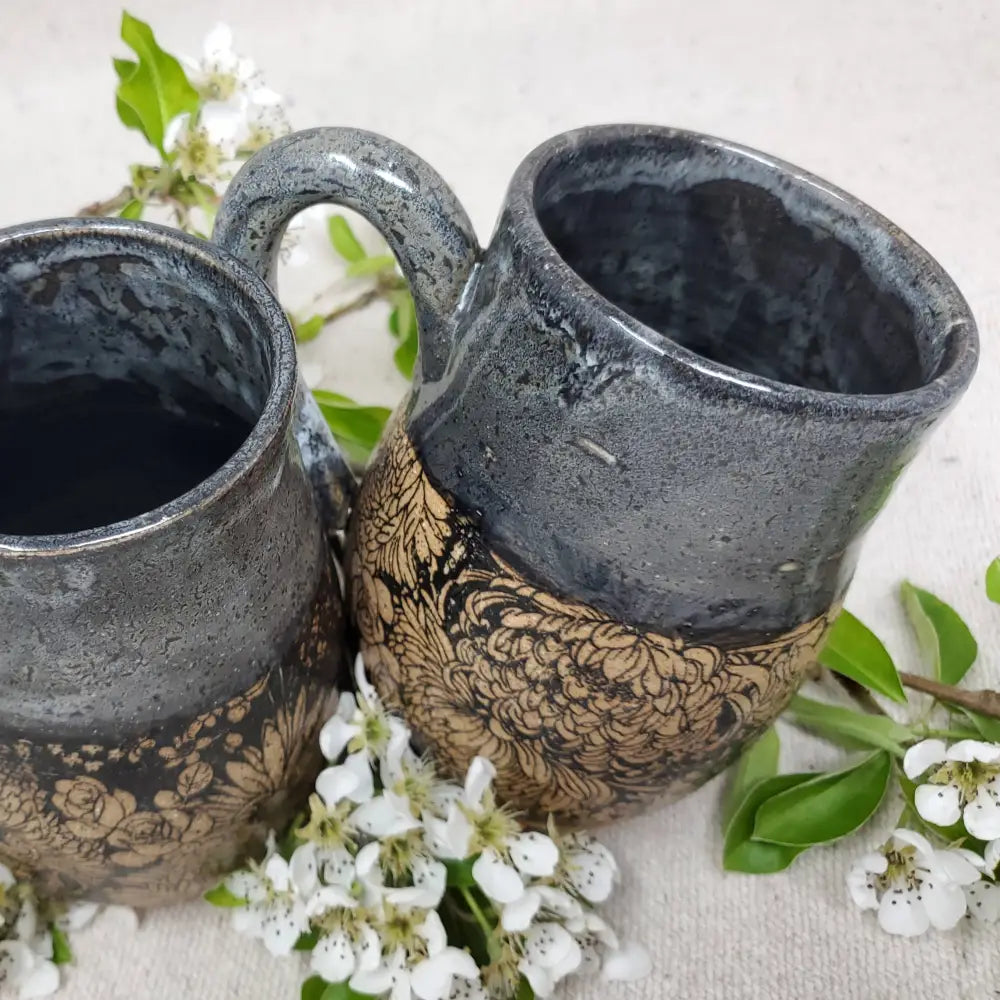 This mug features a handmade, wheel-thrown design. It showcases a distinctive black floral pattern beneath a black glaze accented with a subtle grey-blue overlay