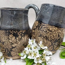 This mug features a handmade, wheel-thrown design. It showcases a distinctive black floral pattern beneath a black glaze accented with a subtle grey-blue overlay