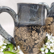This mug features a handmade, wheel-thrown design. It showcases a distinctive black floral pattern beneath a black glaze accented with a subtle grey-blue overlay