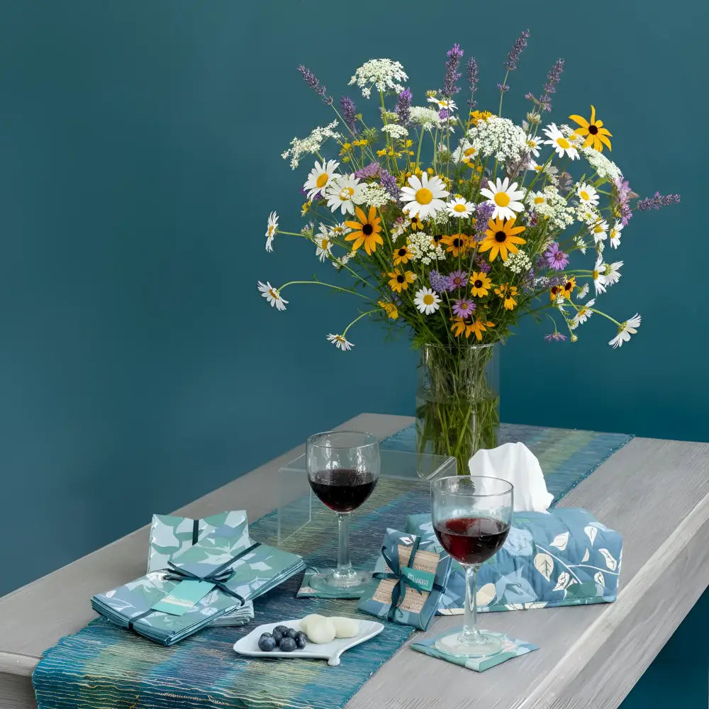 A vibrant wildflower bouquet with daisies sunflowers and lavender in a clear vase complements fair trade blue and green floral napkins set