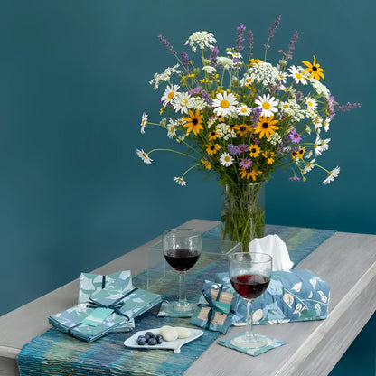 A vibrant wildflower bouquet with daisies sunflowers and lavender in a clear vase complements fair trade blue and green floral napkins set