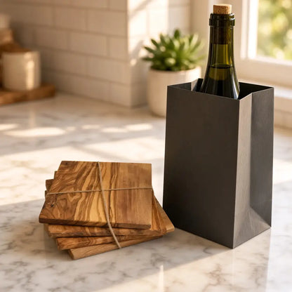 Dark gray paper wine bottle carrier holding green glass bottle with cork next to olive wood four square coasters handcrafted