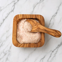 A square olive wood salt bowl with matching wood salt spoon filled with pink Himalayan salt natural grain patterns