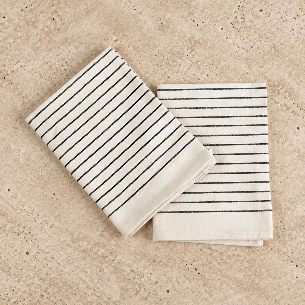 Two neatly folded white organic cotton tea towels with thin black stripes from a wearable kitchen towel set