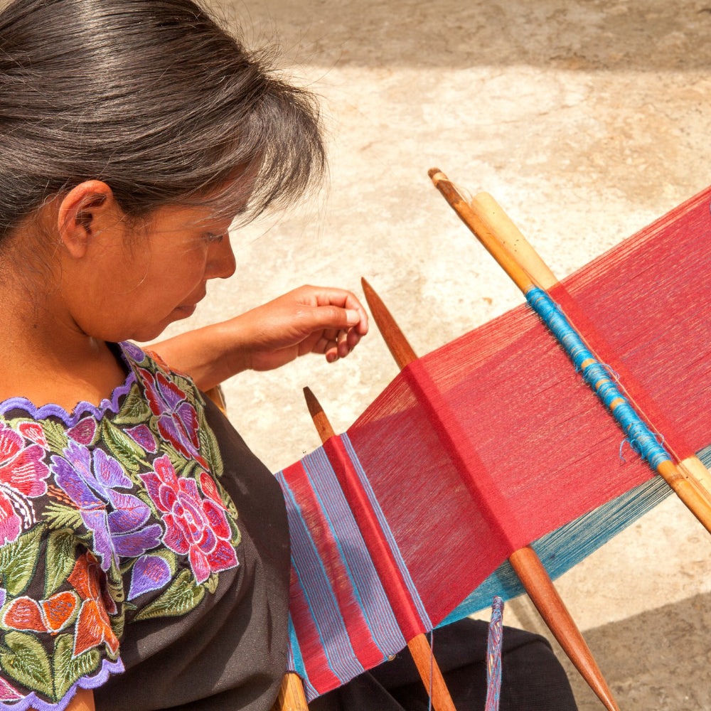 A woman in a black blouse with colorful floral embroidery sits beside a handloom, weaving red, blue, and purple fabric with wooden spindles in an outdoor setting.
