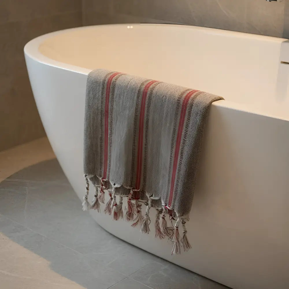 Gray turkish hand towel with red stripes and tassels draped over white bathtub premium cotton hand towels