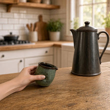 A hand holds a small green divot handmade pitcher with dark rim perfect for pouring cream without awkward grip