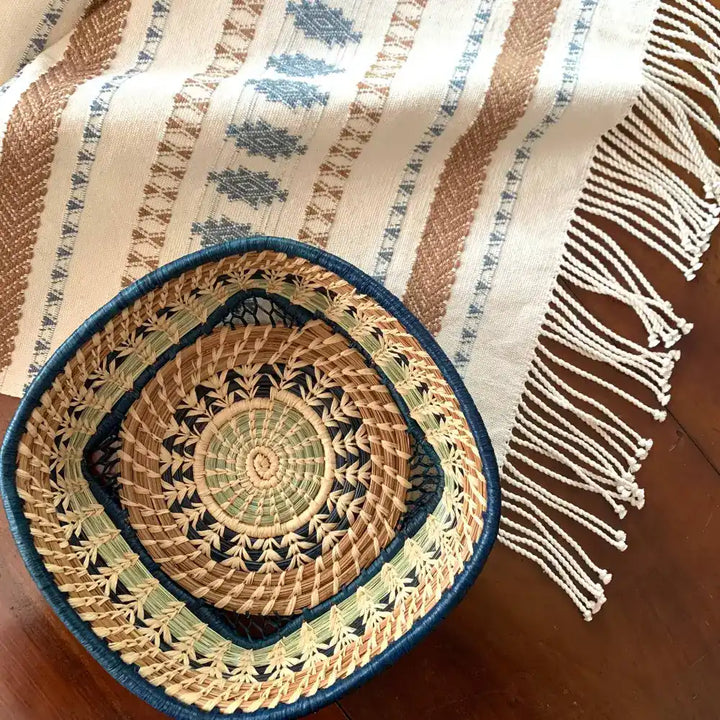 A basket featuring a variety of delicate raffia stitches on coils of pine needles and pajón, a native grass that grows in the highlands of Guatemala. A central circle transitions to a square basket. Indigo blue raffia and pine needles accent the lovely design