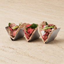 Three stainless steel upright taco racks holding grilled meat and fresh veggie filled tortillas for easy serving
