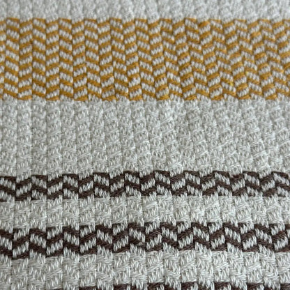 Turkish cotton blanket with a natural, cream colored cotton base woven in an understated modern herringbone pattern with stripes and pom pom edging.