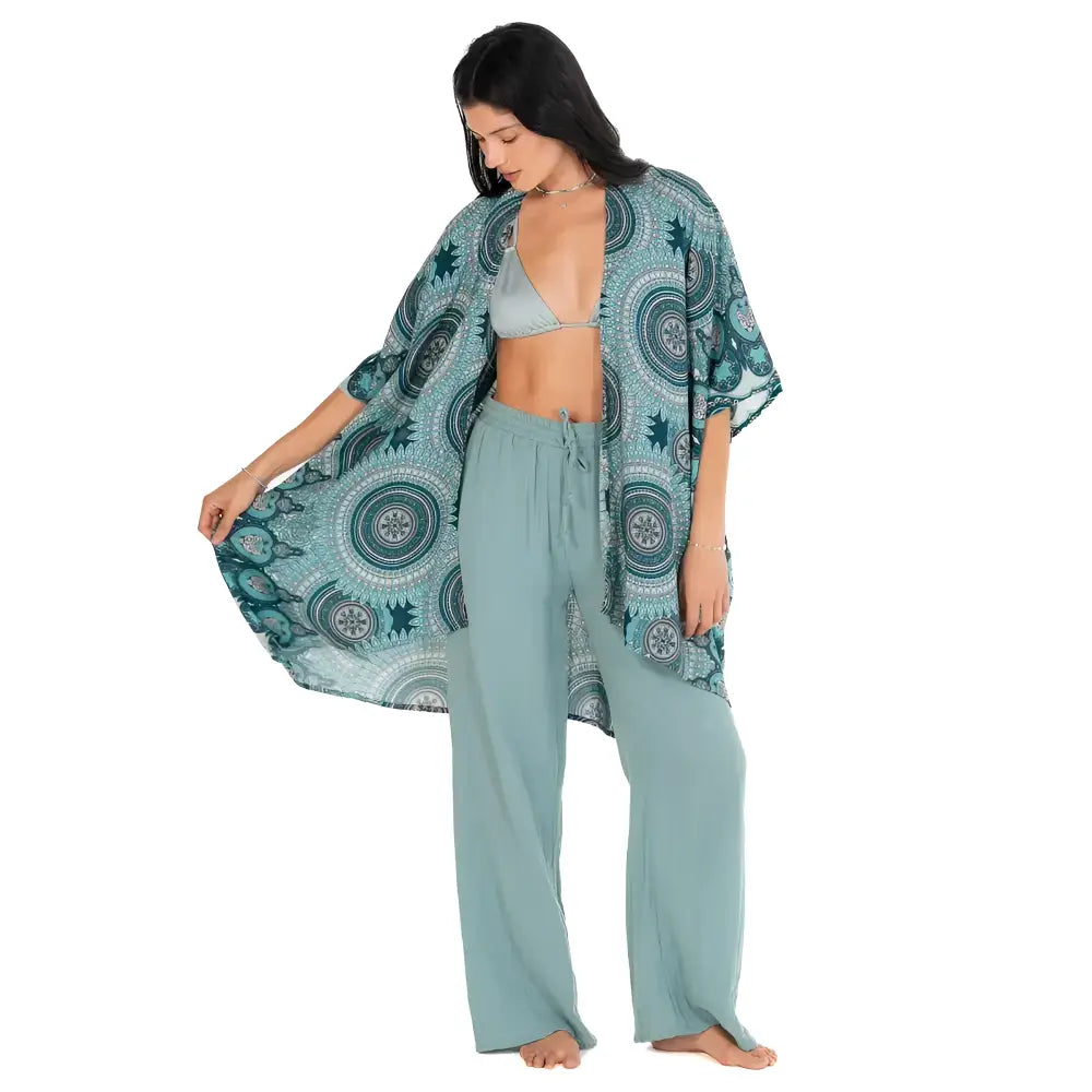 A flowing teal mandala kimono cover-up with circular patterns worn over a light blue bikini top and wide-leg pants