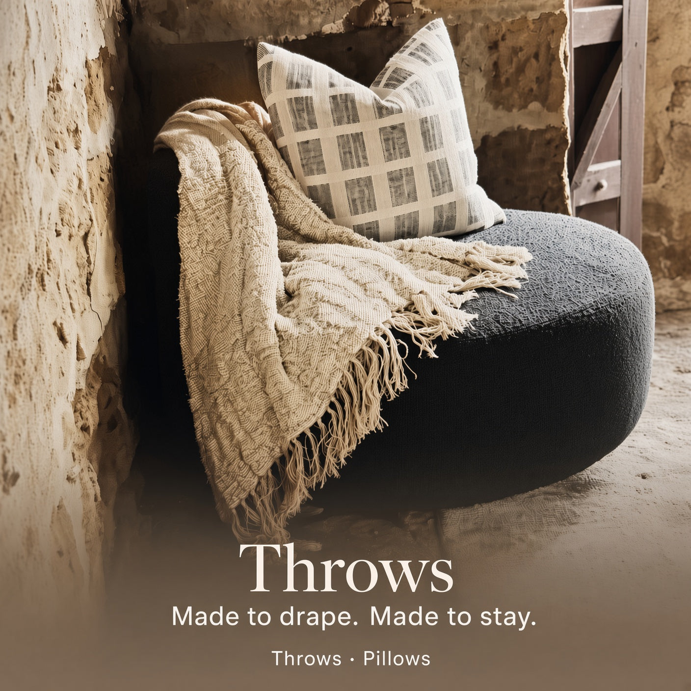 A styled interior scene showing a dark round pouf or bench with a cream fringed throw draped over it and a patterned pillow resting on top. Text at the bottom reads 'Throws Made to drape.
