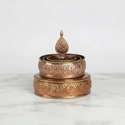 A copper-colored traditional tibetan buddhist offering mandala set featuring an intricately engraved prayer wheel with finial