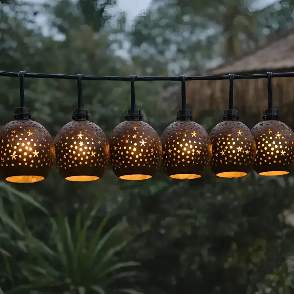 A string of warm-toned spherical outdoor lanterns with hand-carved coconut shades featuring star and dot cutouts