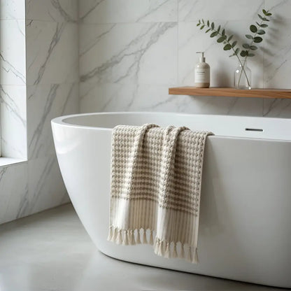 Beige turkish cotton dotted terry long hand towel with tassels draped over a white bathtub