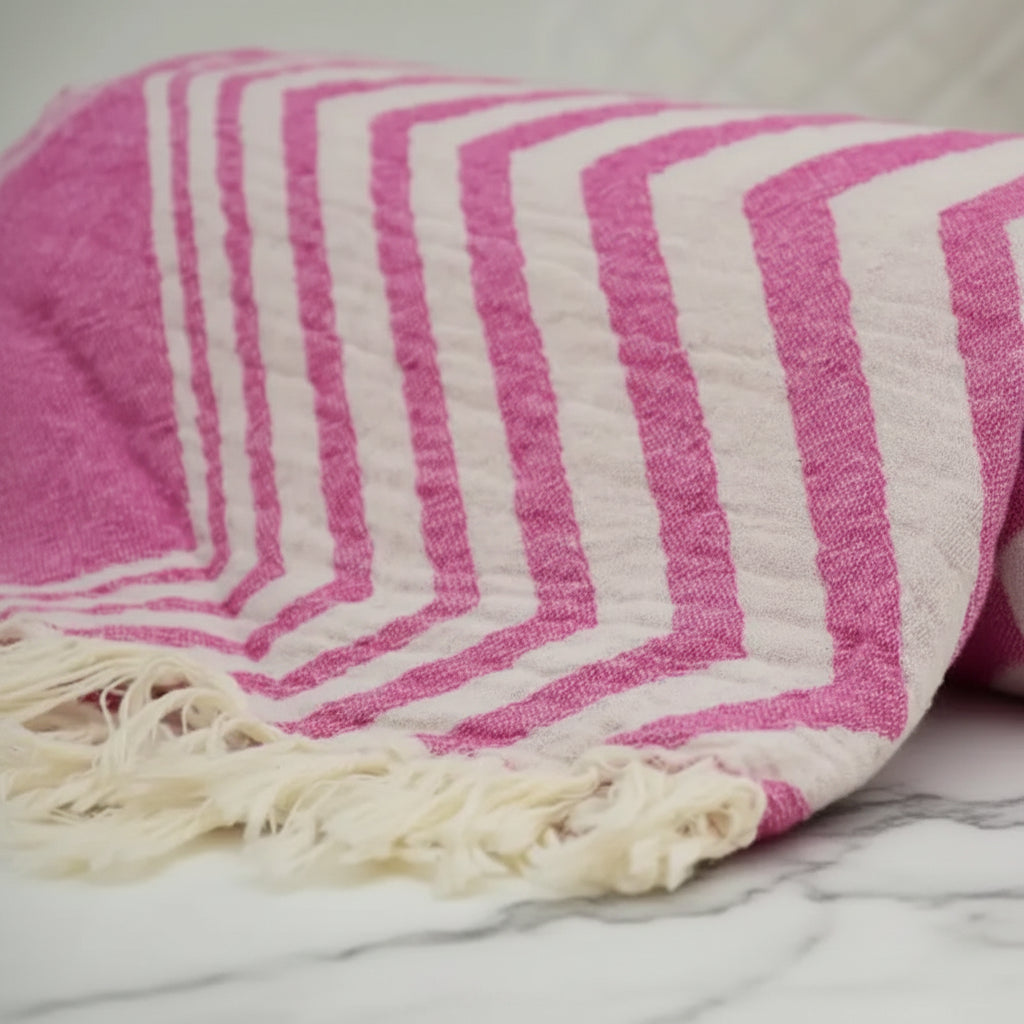 A rolled pink and white striped zigzag pattern peshtemal towel with textured weave and fringed edges