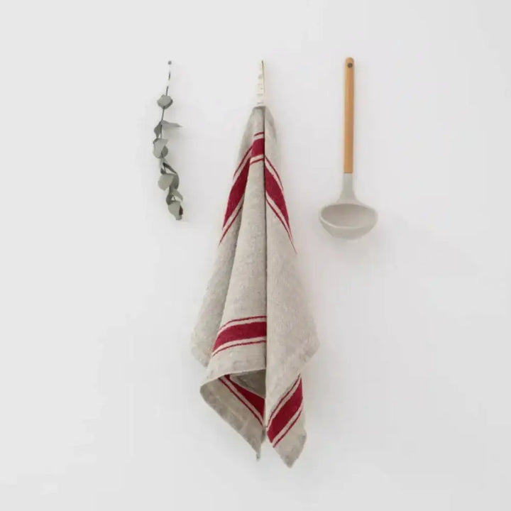 A natural color kitchen towel with red stripes hanging on a hook next to kitchen utensils.