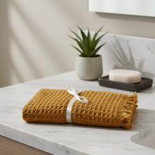 A mustard-yellow waffle soft turkish cotton hand towel with fringed edges rolled and tied with a white ribbon