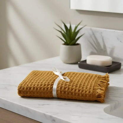 A mustard-yellow waffle soft turkish cotton hand towel with fringed edges rolled and tied with a white ribbon