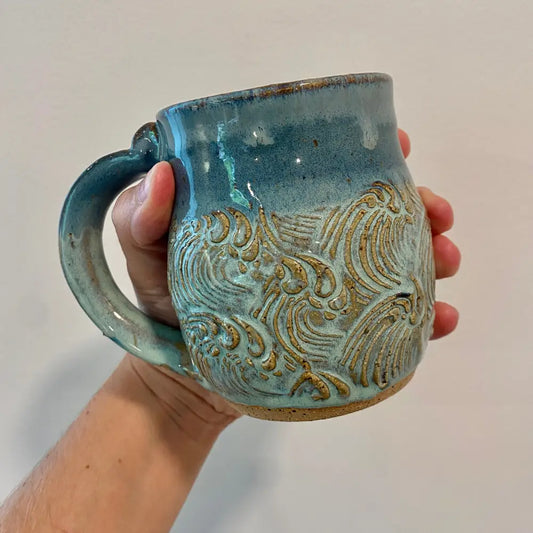 This handmade pottery mug features a textured sea-foam wave design with ocean blue rim.