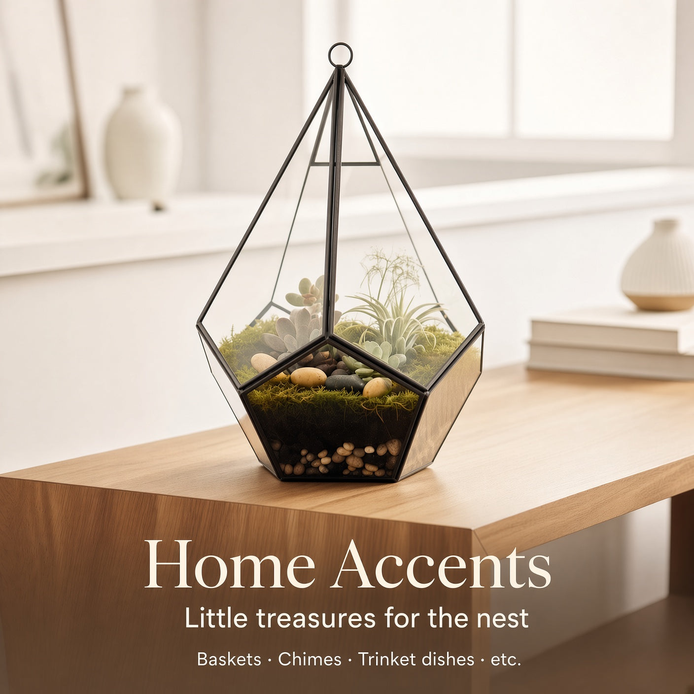 A geometric glass terrarium with a black metal frame sits on a light wood table in a softly lit home interior. The terrarium contains moss, small plants, and pebbles.