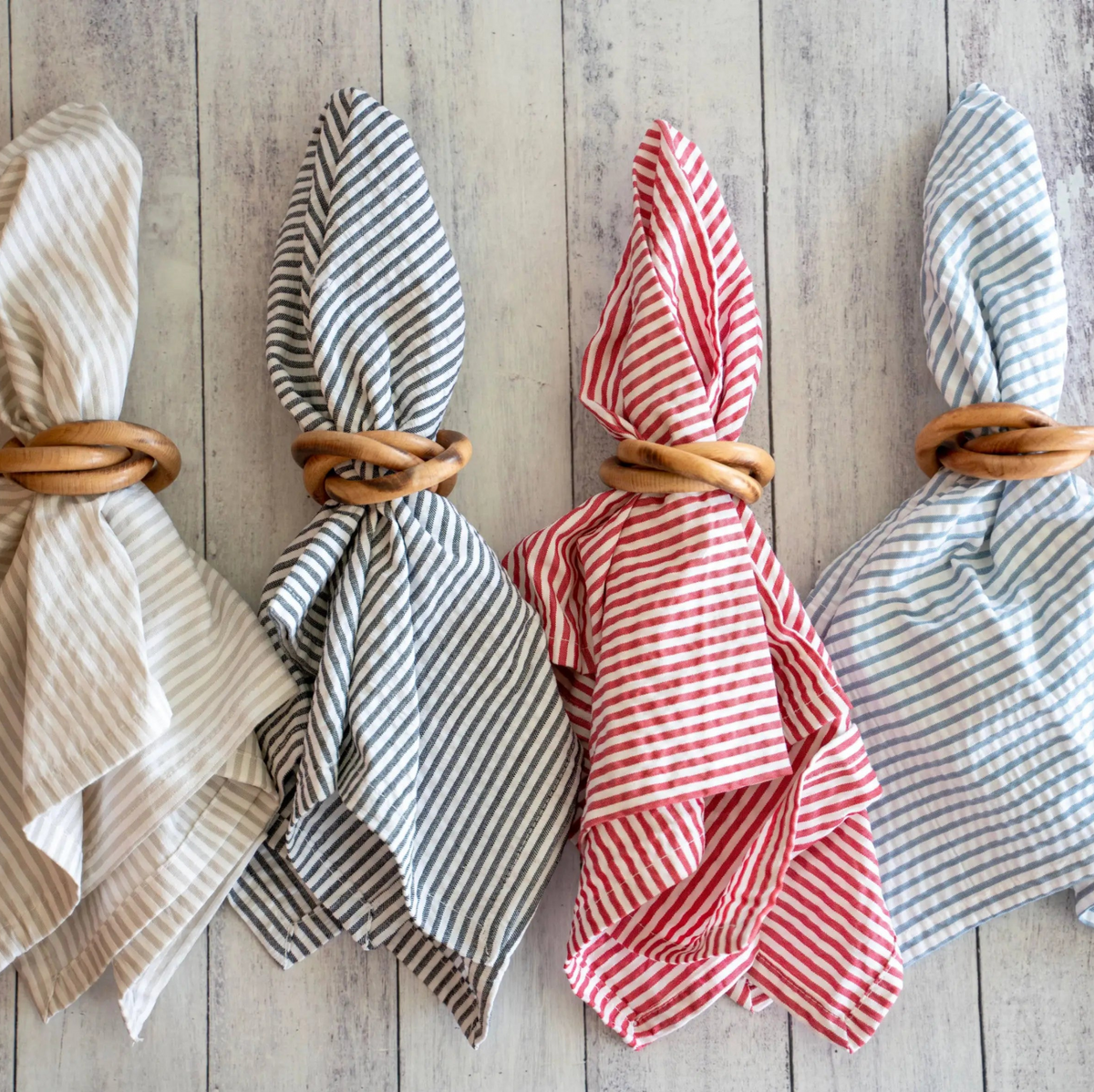 Four napkins with striped designs and wooden napkin rings in a row on a whitewashed wooden board.