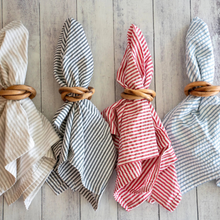 Four napkins with striped designs and wooden napkin rings in a row on a whitewashed wooden board.